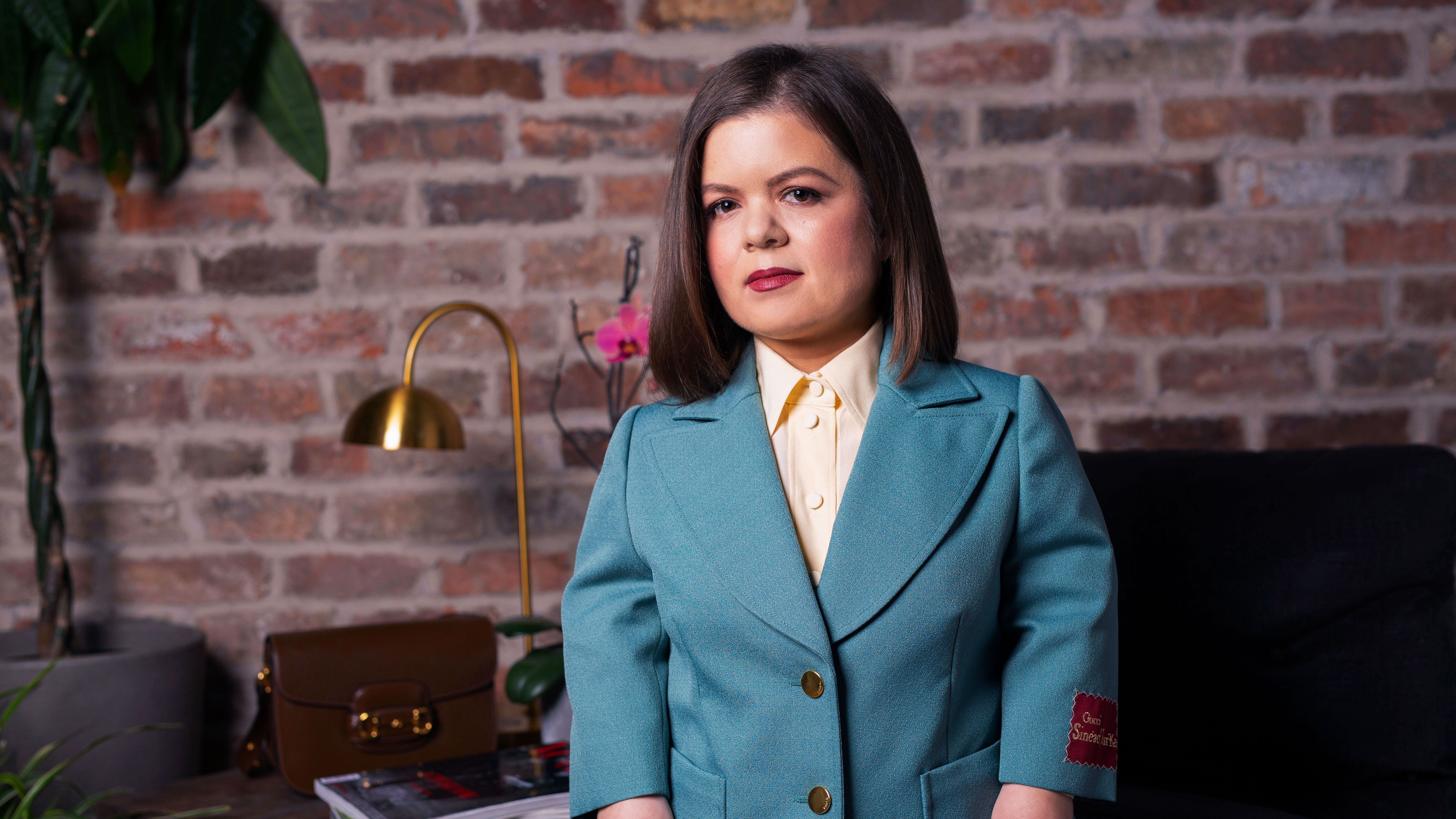 Sinead Burke, a white, visibly Disabled woman, stands in front of a brick wall in a teal Gucci suit.
