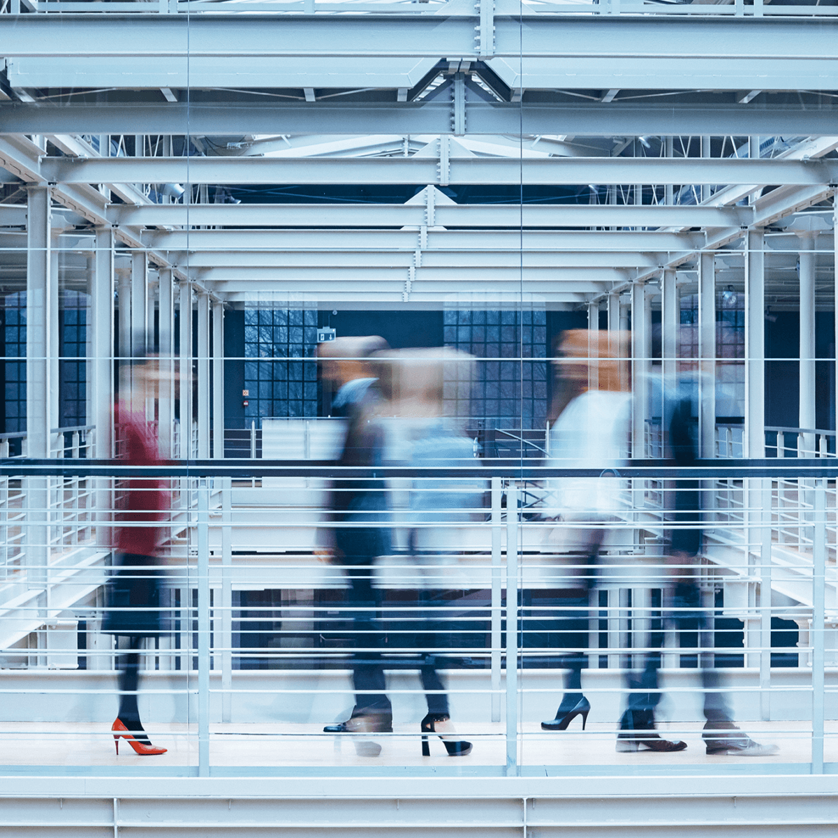 An image of an office with blurred figures in suits walking along a walkway. Three of the figures wear high heels.