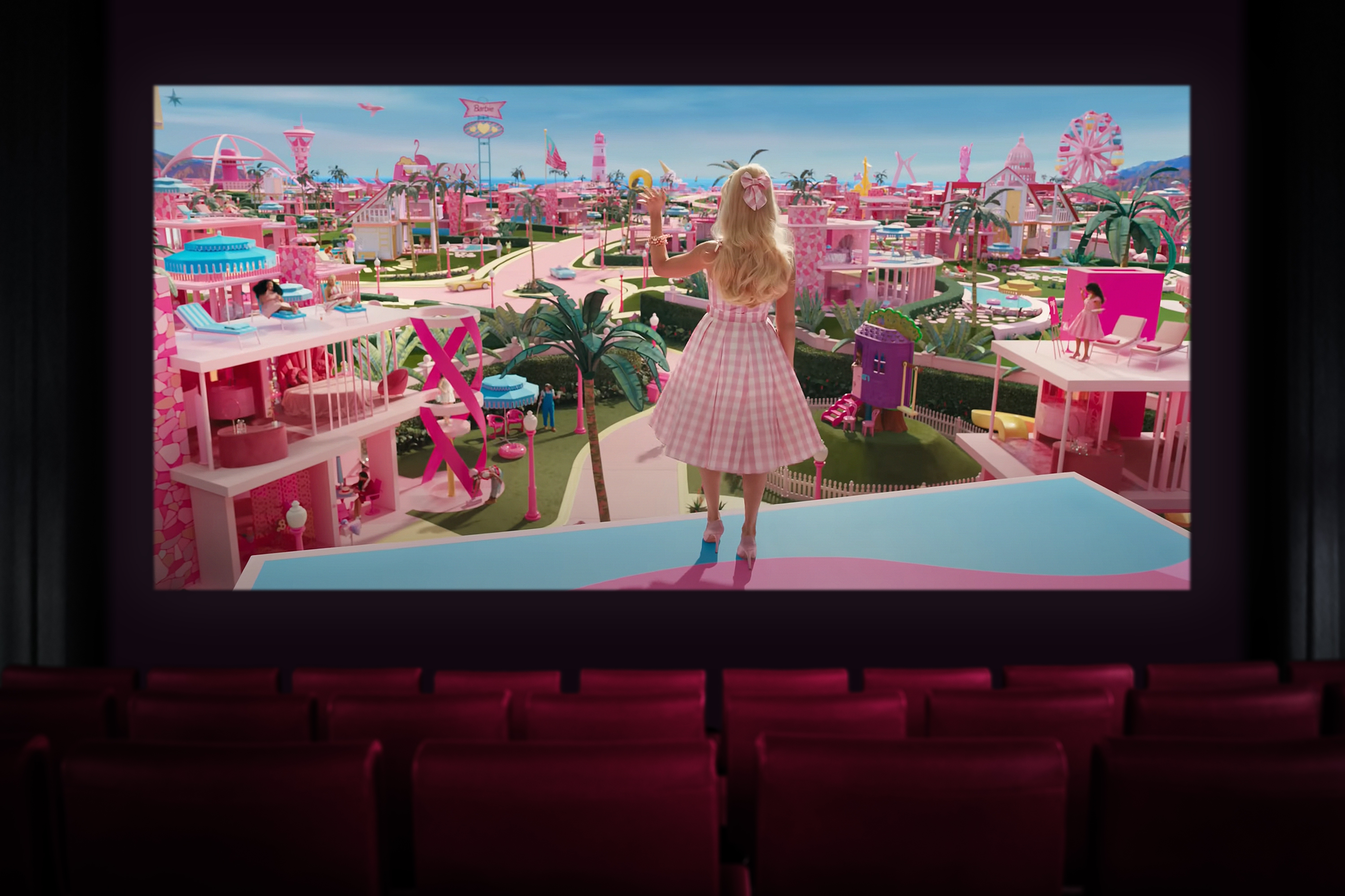 cinema seats in front of a screen showing the Barbie movie. In it, Barbie is stood on top of a building, waving at other Barbies.
